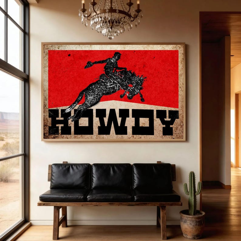May include: A framed art print featuring a rodeo scene with a cowboy riding a bucking bronco against a red background. The word "HOWDY" is displayed in bold, black letters. The artwork is displayed above a black leather bench.