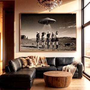 May include: A large rectangular wall art print in a wooden frame featuring a black and white desert landscape with five people in cowboy hats standing in a row, reaching toward a hovering UFO emitting a beam of light, with a small cow nearby.