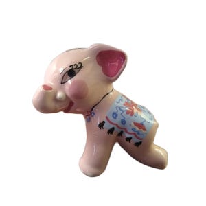 May include: A pink ceramic elephant figurine with a blue and floral patterned blanket. The elephant has pink ears, cheeks, and a trunk. Black details include eyelashes and small feet.