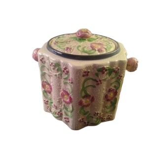 Peut inclure: Un pot hexagonal en céramique avec couvercle, doté d'une base couleur crème à la surface texturée. L'extérieur est orné de fleurs roses en relief peintes à la main, de vignes vertes et de petits accents rouges. Il possède deux anses latérales.
