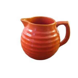 May include: A ceramic jug in a bright orange hue, featuring a rounded form and a handle. The body is adorned with horizontal ridges. It has a spout for pouring and is suitable for serving drinks. The jug is likely made of earthenware.