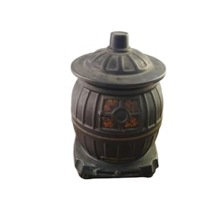 May include: A dark gray, vintage-style decorative container shaped like a small stove. The container has a domed lid and a barrel-like body with vertical lines. A small, round window with orange and red floral accents is visible on the front.