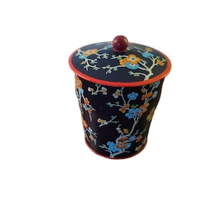 May include: A black decorative container with a lid, featuring a floral pattern in orange, blue, and white. The rim and lid edge are red, and the lid has a red knob. The container is cylindrical and appears to be made of a fabric-like material.