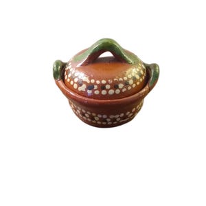 May include: A small, round, terracotta-colored ceramic pot with a lid. The pot is decorated with white polka dots and has a green handle and accents. The lid fits snugly on the pot, and the overall design is rustic and charming.