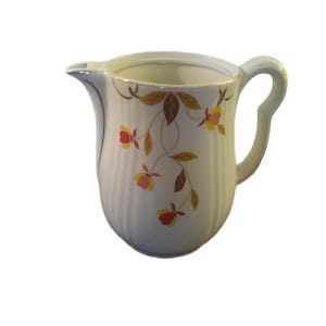 May include: White ceramic creamer with a curved handle and spout. The creamer features a vertical ribbed design and is decorated with a floral pattern of yellow, red, and brown flowers and leaves. A vintage-style serving piece.