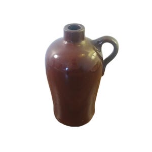 May include: A brown ceramic jug with a handle. The jug has a rounded body and a narrow neck with a small opening. The handle is arched and attached to the side of the jug. The jug is a dark brown color and has a glossy finish.