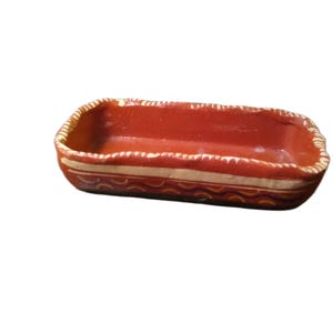 May include: A rectangular terracotta dish with a reddish-brown interior and a textured, uneven rim. The exterior features a cream-colored horizontal band above a wavy line pattern in dark blue and orange, set against a reddish-brown base.