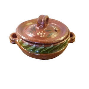 May include: A small, round, terracotta-colored ceramic pot with a lid. The lid has a handle and is decorated with white dots and green leaf designs. The pot has two side handles and green and white painted accents.