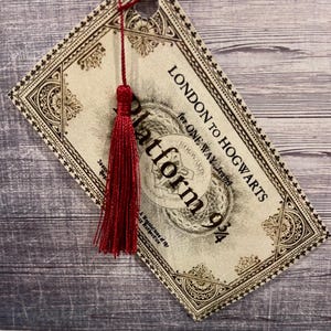 Op de afbeelding: Een ticket in vintage-stijl met de tekst "LONDON TO HOGWARTS" en "Platform 9 3/4". Het ticket is crèmekleurig met decoratieve randen en een rode kwast. De achtergrond is een grijs houten oppervlak.