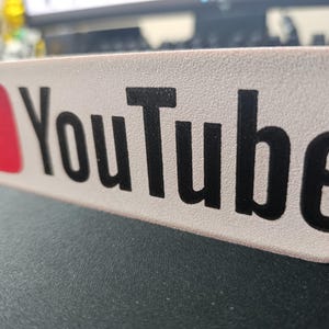 Può includere: Un cartello rettangolare con il logo di YouTube. Il logo presenta un quadrato rosso con un pulsante di riproduzione triangolare bianco e la parola "YouTube" in nero, carattere sans-serif.