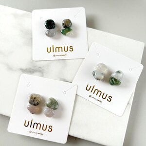 May include: Three pairs of stud earrings featuring two stacked, polished, irregular-shaped stones in shades of green, white, and grey. Each pair is mounted on a white display card printed with the gold text ulmus and an Instagram handle.