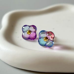 以下が含まれることがあります： 2つの花の形をしたスタッドピアス。各イヤリングは、青、紫、ピンクの色合いの半透明のデザインと、小さな金色の中心部が特徴です。イヤリングは、白い波状の縁のあるトレイに表示されています。