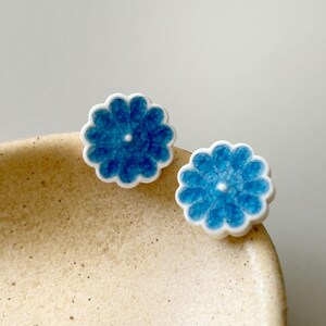 Puede incluir: Un par de pendientes de botón azules con forma de flor y bordes festoneados blancos. Los pendientes tienen un aspecto texturizado, similar a un mosaico, y un pequeño centro blanco. Se muestran sobre una superficie beige.
