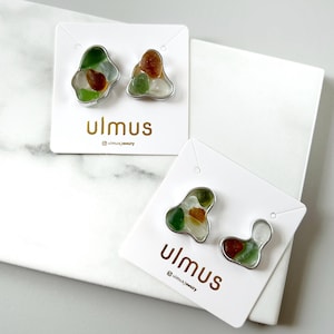 May include: Two pairs of stud earrings with irregular shapes, each featuring a mix of green, brown, and white sea glass set in silver-colored metal. The earrings are displayed on white cards with the brand name "ulmus".