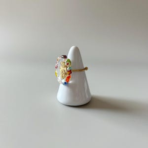 May include: A gold ring with a clear, oval-shaped centerpiece. The centerpiece features a gold design and is surrounded by colorful glass beads. The ring is displayed on a white, cone-shaped stand.