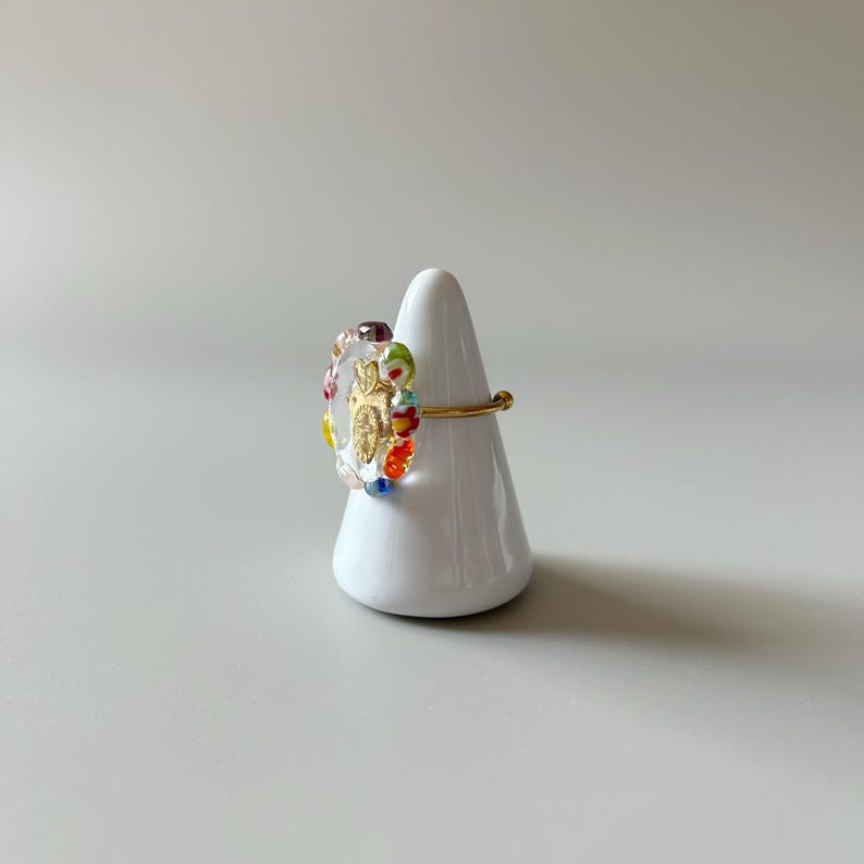 May include: A gold ring with a clear, oval-shaped centerpiece. The centerpiece features a gold design and is surrounded by colorful glass beads. The ring is displayed on a white, cone-shaped stand.