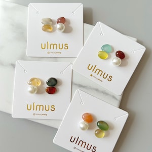 May include: Four pairs of gemstone stud earrings on white cards. Each card features the gold text "ulmus" and the brand's social media handle. The earrings showcase a variety of colors, including yellow, orange, green, red, and white.