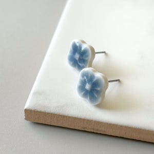 May include: A pair of light blue flower-shaped stud earrings. The earrings have a white border and a light blue centre. The earrings are on a white surface.