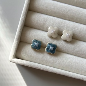 Puede incluir: Cuatro pares de pendientes de botón con forma de trébol. Dos pares son blancos con un centro de perla, y dos pares son verde azulado con un centro de perla. Los pendientes se muestran en una caja de joyería blanca.