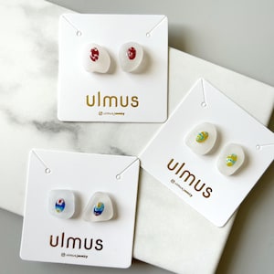 Puede incluir: Tres pares de pendientes de botón blancos con diseños coloridos se exhiben en tarjetas blancas. Los pendientes tienen formas irregulares y presentan detalles en rojo, azul y amarillo. Las tarjetas tienen el nombre de la marca "ulmus" impreso en dorado.