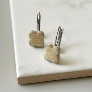 Puede incluir: Un par de pendientes con diseño de trébol de cuatro hojas. Los pendientes son de color beige claro con cierres de palanca plateados. Los pendientes están sobre una superficie blanca.