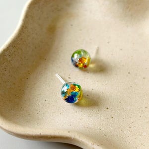 Puede incluir: Dos pendientes de botón con un diseño esférico transparente. Cada pendiente presenta un patrón abstracto colorido de azul, amarillo, naranja y verde dentro de la esfera transparente. Los pendientes están sobre un fondo neutro blanquecino.