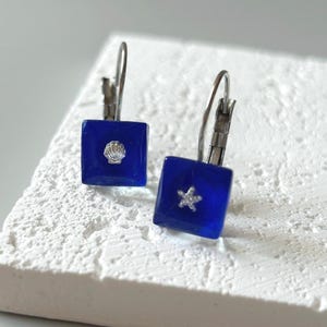 May include: A pair of square, cobalt blue earrings with silver-tone lever-back closures. One earring features a seashell, the other a starfish, both in silver. The earrings are displayed on a textured white surface.