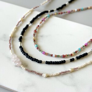 May include: Close-up of three beaded necklaces. One necklace features black beads, another has a mix of colourful beads, and the third has pink and white beads. The necklaces are on a white surface.