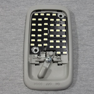 May include: A grey interior car light fitting with a rectangular array of small, yellow LED lights. The fitting has a grey plastic housing with a switch labelled "ON OFF DOOR". A metal screw and a grey lever are also visible.