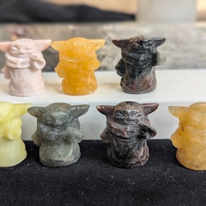 Op de afbeelding: Zeven kleine, gebeeldhouwde beeldjes van een personage met grote oren en een gewaad. De beeldjes zijn in verschillende kleuren, waaronder roze, geel, grijs en bruin. De figuren staan rechtop.