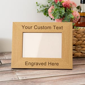 May include: A rectangular, light-colored wooden picture frame with the text "Your Custom Text" and "Engraved Here" engraved on the frame. The frame is empty, ready for a custom photo. The frame is sitting on a wooden surface.