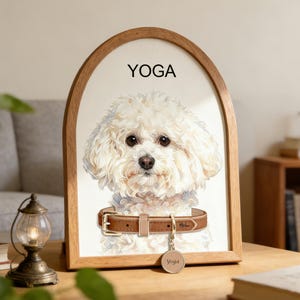 以下が含まれることがあります： 茶色の革の首輪と「Yoga」と書かれたタグを付けた、ふわふわの白い犬が描かれた額入りのプリント。犬の上には「YOGA」の文字が印刷されています。木製のフレームは木製の表面に置かれています。