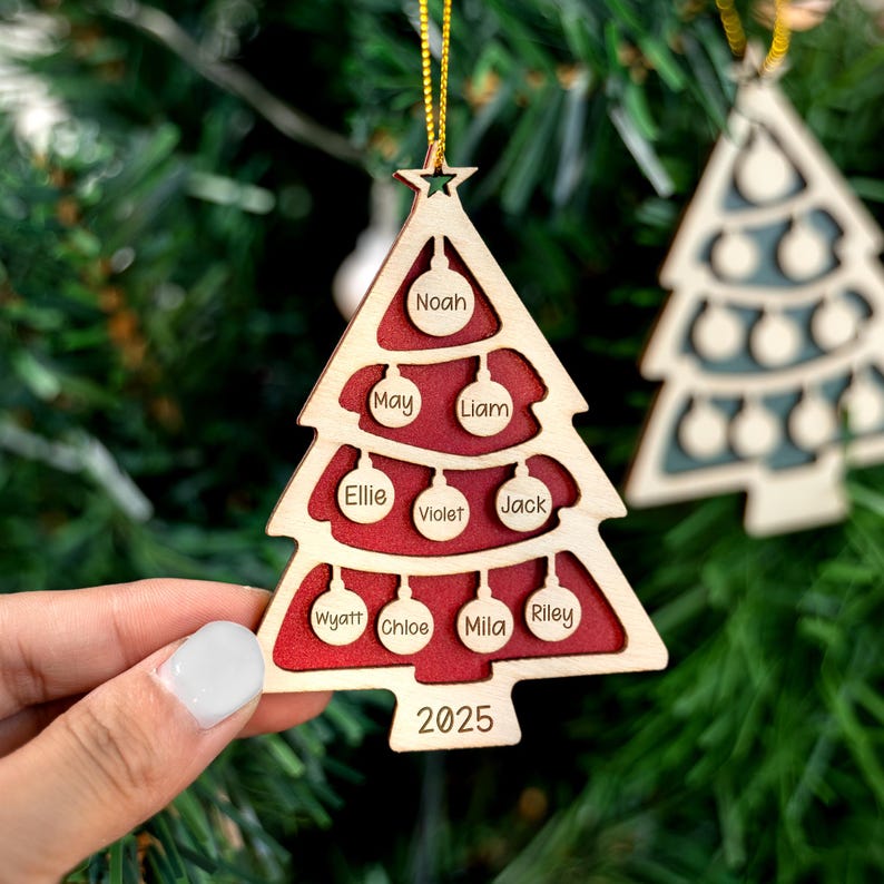 May include: Wooden Christmas tree ornament with a red interior and the names Noah, May, Liam, Ellie, Violet, Jack, Wyatt, Chloe, Mila, and Riley. The year 2025 is engraved at the bottom. The ornament is hanging on a gold string.