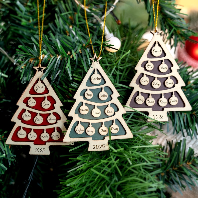 May include: Three wooden Christmas tree decorations, each shaped like a tree with a star and gold hanging string. The ornaments are personalised with names and the year 2025. The trees are red, teal, and grey, with names in small circles.