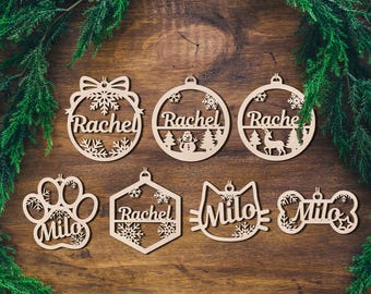 May include: Wooden Christmas ornaments in various shapes, including a paw print, cat face, and bone, with names like "Milo" and "Rachel" cut out. Snowflake and winter scene designs are incorporated. The ornaments are displayed on a wooden surface.