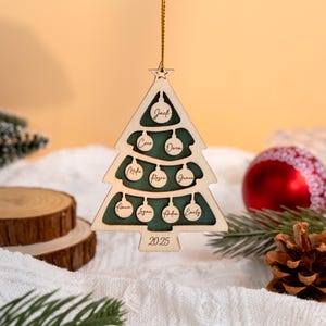 May include: Wooden Christmas tree decoration in the shape of a tree, with a green interior and names including Jack, Coco, and Owen. The year 2025 is engraved at the bottom. It hangs from a gold string.