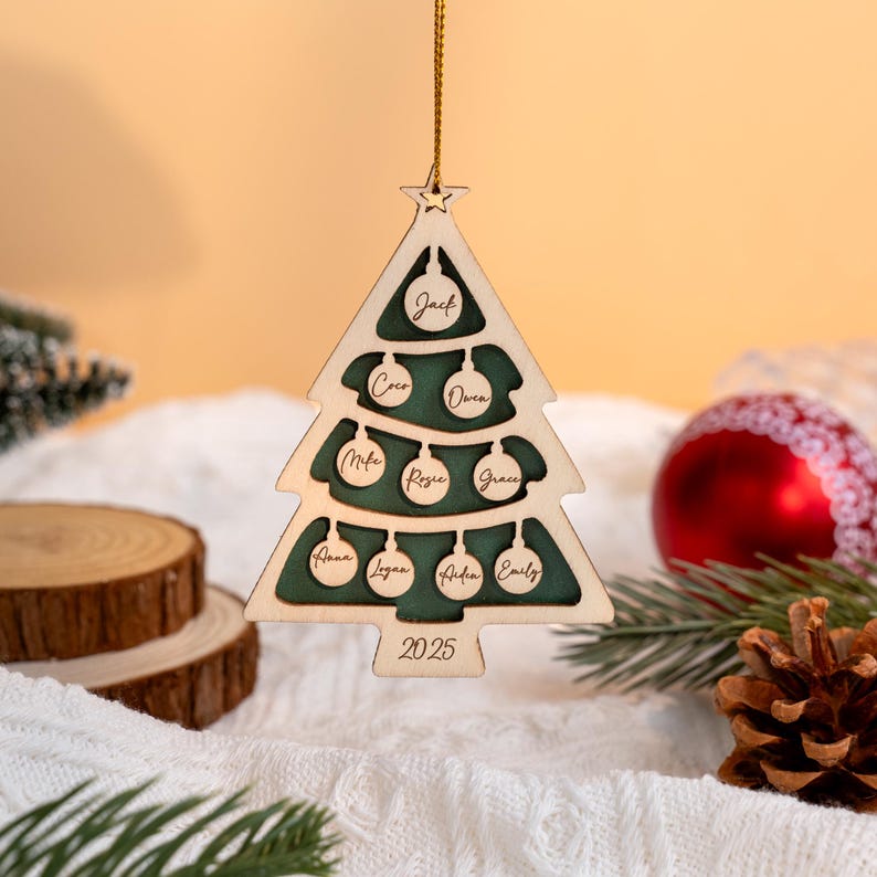 May include: Wooden Christmas tree decoration in the shape of a tree, with a green interior and names including Jack, Coco, and Owen. The year 2025 is engraved at the bottom. It hangs from a gold string.