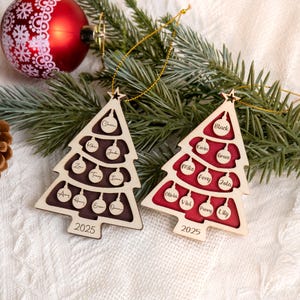 May include: Two wooden Christmas tree ornaments with personalized names and the year 2025. One tree has a dark brown interior, the other has a red interior. Both are hanging on a gold string, with a red ornament and pine branches in the background.