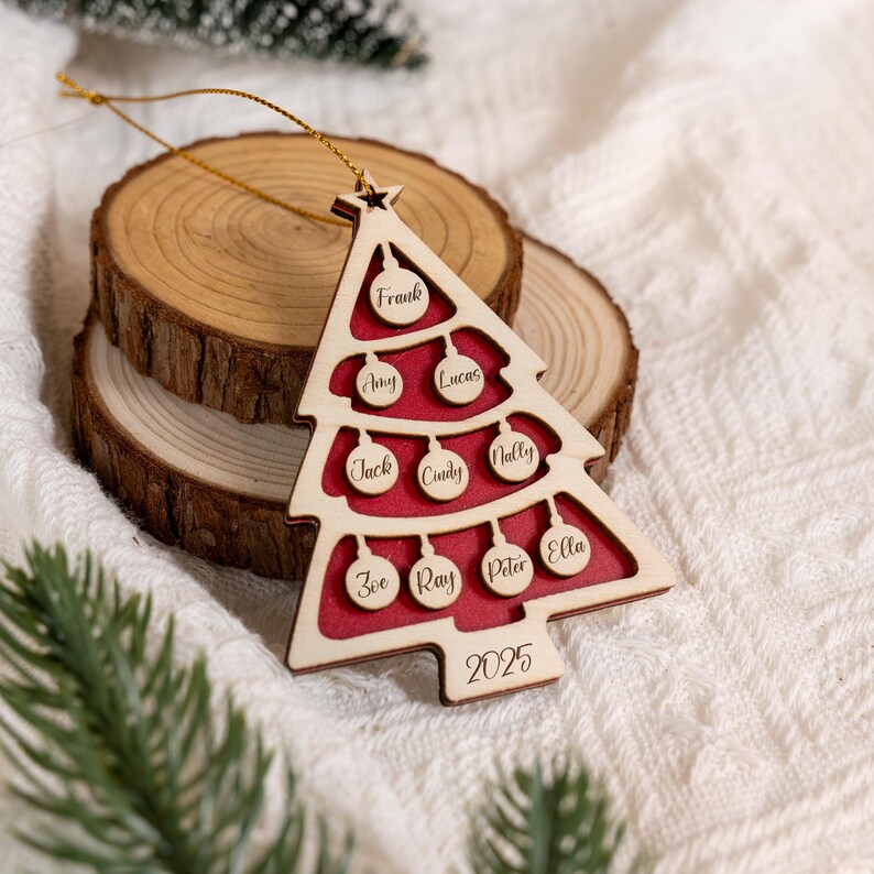 May include: A wooden Christmas tree decoration with a red interior and names engraved on the baubles. The decoration is shaped like a Christmas tree and has the year 2025 engraved at the bottom. A gold string is attached for hanging.