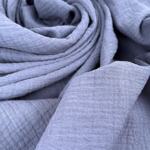 May include: A soft, periwinkle blue fabric featuring a distinct crinkled, double-gauze texture. The material is draped in loose, flowing folds, highlighting the fine, puckered surface pattern and the consistent, muted blue color throughout the textile.