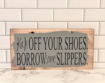 Peut inclure: Une pancarte en bois peinte en gris avec une inscription noire qui dit "Kick off your shoes. Borrow some slippers."