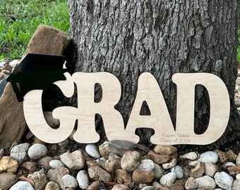 May include: Wooden GRAD sign with a black graduation cap on top. The sign is personalized with the name "Naomi Noble" and the text "Class of 2024".