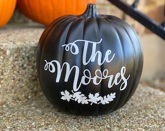 Puede incluir: Una calabaza decorativa negra con el texto blanco "The Moores". La calabaza tiene un tallo y un diseño de hojas en la parte inferior. Otras calabazas son visibles en el fondo.