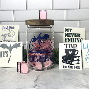 May include: A clear glass jar with a wooden lid, filled with pink petals and a blue dragon design. Surrounding the jar are small white cards with black and blue text, and pink miniature book-shaped decorations. The theme is books.