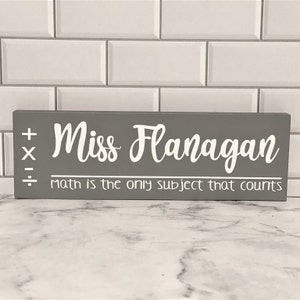 May include: Gray wooden sign with white text that reads "Miss Flanagan, math is the only subject that counts". The sign also has white plus, minus, multiplication, and division symbols.
