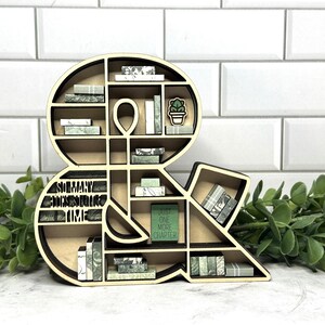 May include: A wooden ampersand-shaped bookshelf with multiple shelves. The shelves are filled with miniature books in shades of green and white. The text "SO MANY BOOKS SO LITTLE TIME" is carved into the design. A small green book reads "JUST ONE MORE CHAPTER".