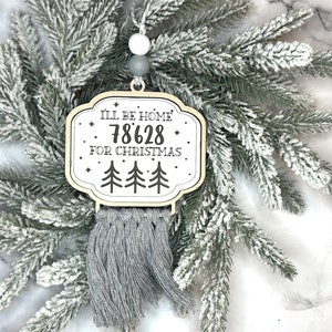 May include: A wooden Christmas ornament with the text "I'll be home 78628 for Christmas" and three trees. The ornament has a gray macrame fringe and is hanging on a frosted evergreen branch.