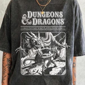 Peut inclure: T-shirt gris foncé avec l'inscription "DUNGEONS & DRAGONS" au-dessus d'une illustration en noir et blanc d'un dragon et d'un guerrier. Le t-shirt a un aspect vintage et délavé.