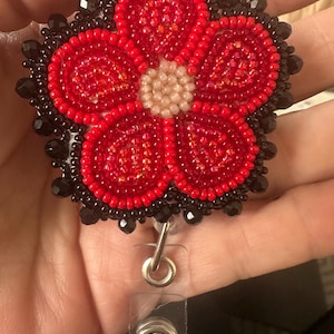 May include: A circular badge reel featuring a five-petaled flower design crafted from red and black seed beads. The flower centre is composed of pale pink beads, while the outer edge is accented with larger black faceted beads and a clear strap.