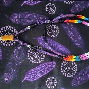 May include: A long, tubular beaded necklace featuring a gradient pattern of purple, black, white, orange, yellow, blue, and pink glass beads. The necklace includes a tan leather connector piece at one end and a white cord loop at the other end.
